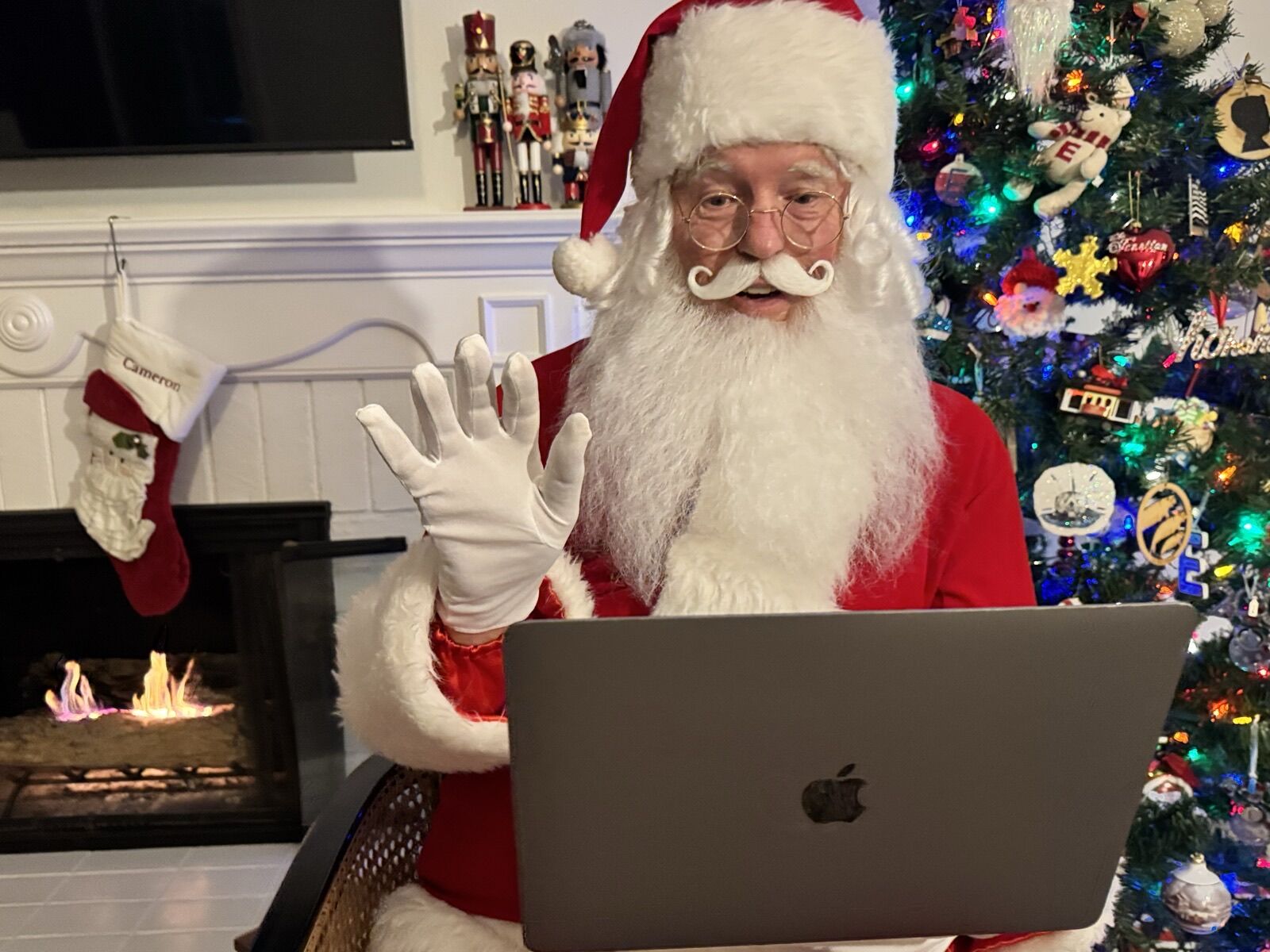Santa Claus waving hello on a laptop video call beside a fireplace